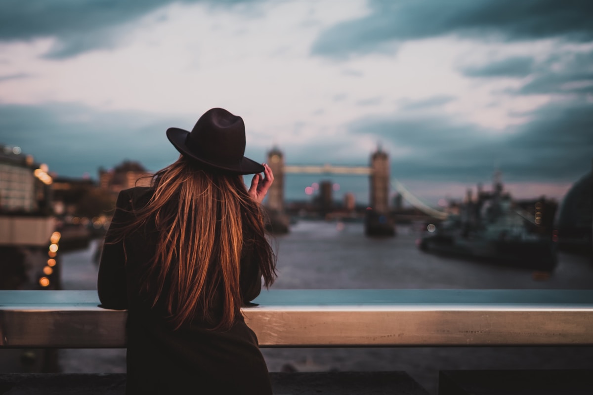 selective focus photo of back of woman wearing fedora hat with elbows on railings facing bridge - stay safe in London, plan trip to London