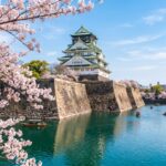 Osaka Castle surrounded by blooming cherry blossoms and a serene river, capturing the essence of springtime beauty in Japan.