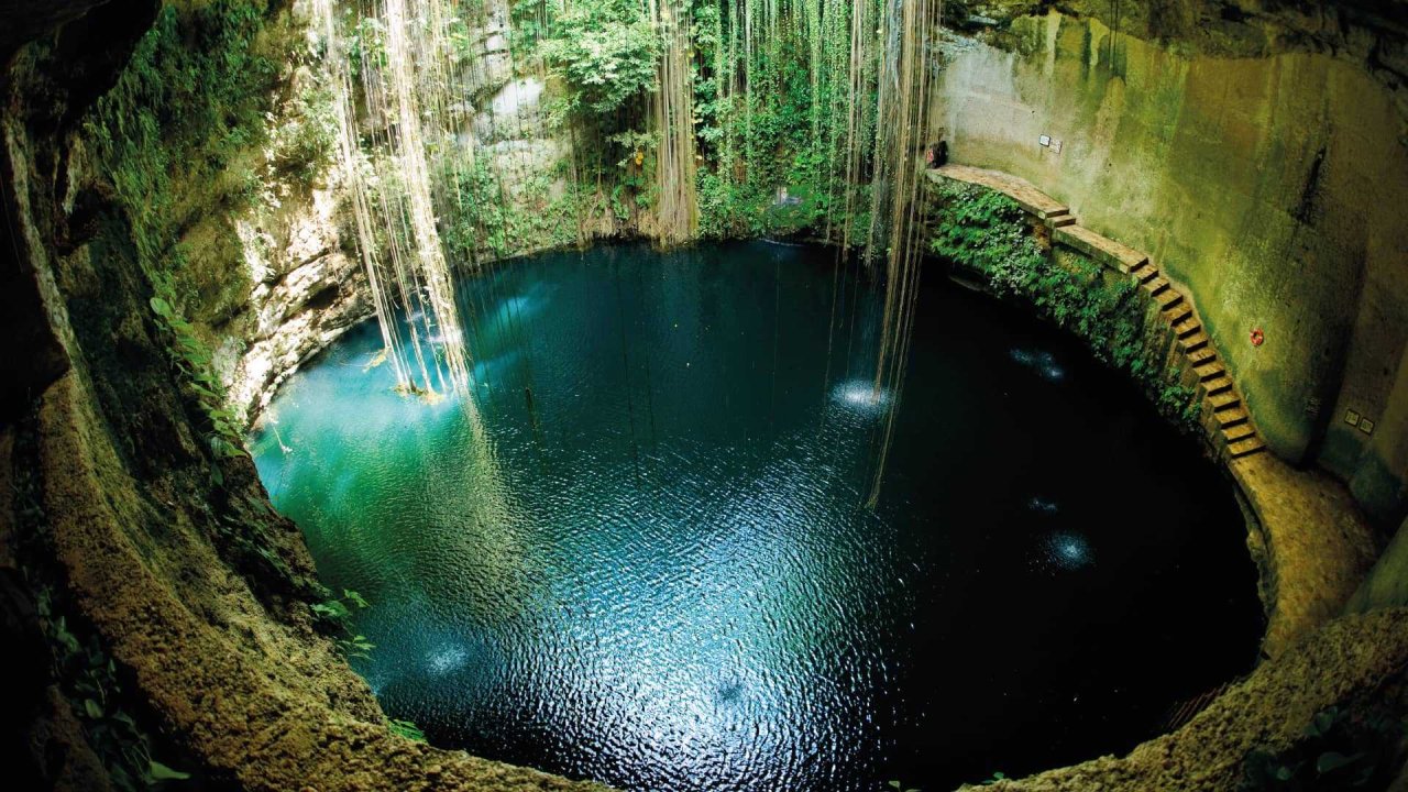 Cenote in Mexico with crystal-clear waters surrounded by lush greenery, featuring hanging vines and stone steps.