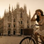 Kannaya Nareswari in a sundress riding a bike near Milan's stunning cathedral during sunset, embodying joyful urban exploration.