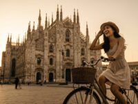 Kannaya Nareswari in a sundress riding a bike near Milan's stunning cathedral during sunset, embodying joyful urban exploration.
