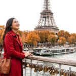 Woman in a red coat smiles near the Eiffel Tower, surrounded by autumn foliage and love locks along the riverbank.