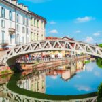 Charming canal scene in Milan featuring a unique bridge, bustling shops, and vibrant reflections by the water's edge.