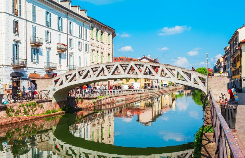 Charming canal scene in Milan featuring a unique bridge, bustling shops, and vibrant reflections by the water's edge.