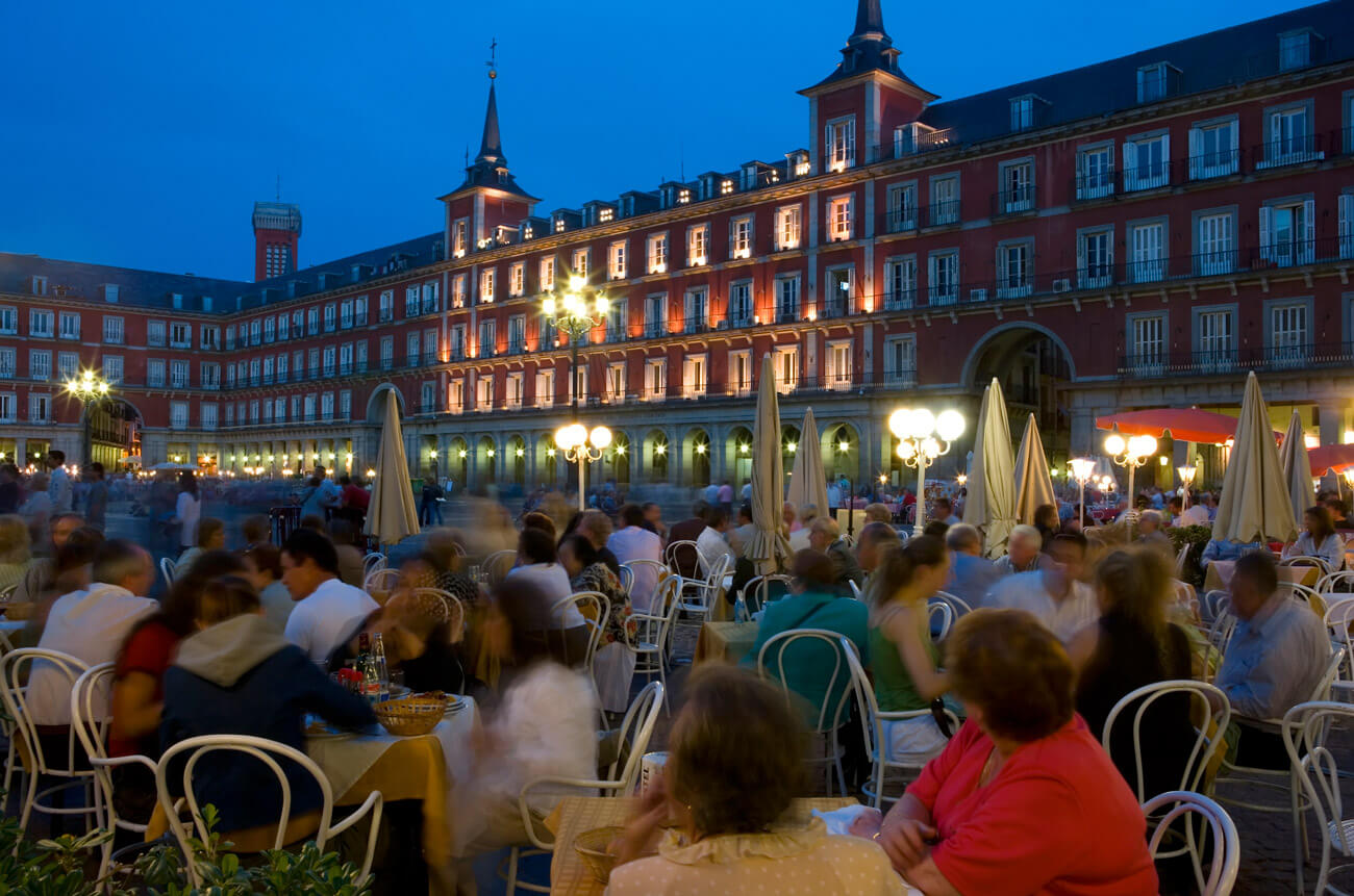 Vibrant outdoor dining scene in Madrid's Plaza Mayor, with lively crowds enjoying the evening atmosphere under warm lights.