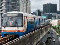 BTS Skytrain traveling on an elevated track amidst modern buildings in a bustling urban setting of Bangkok, Thailand.
