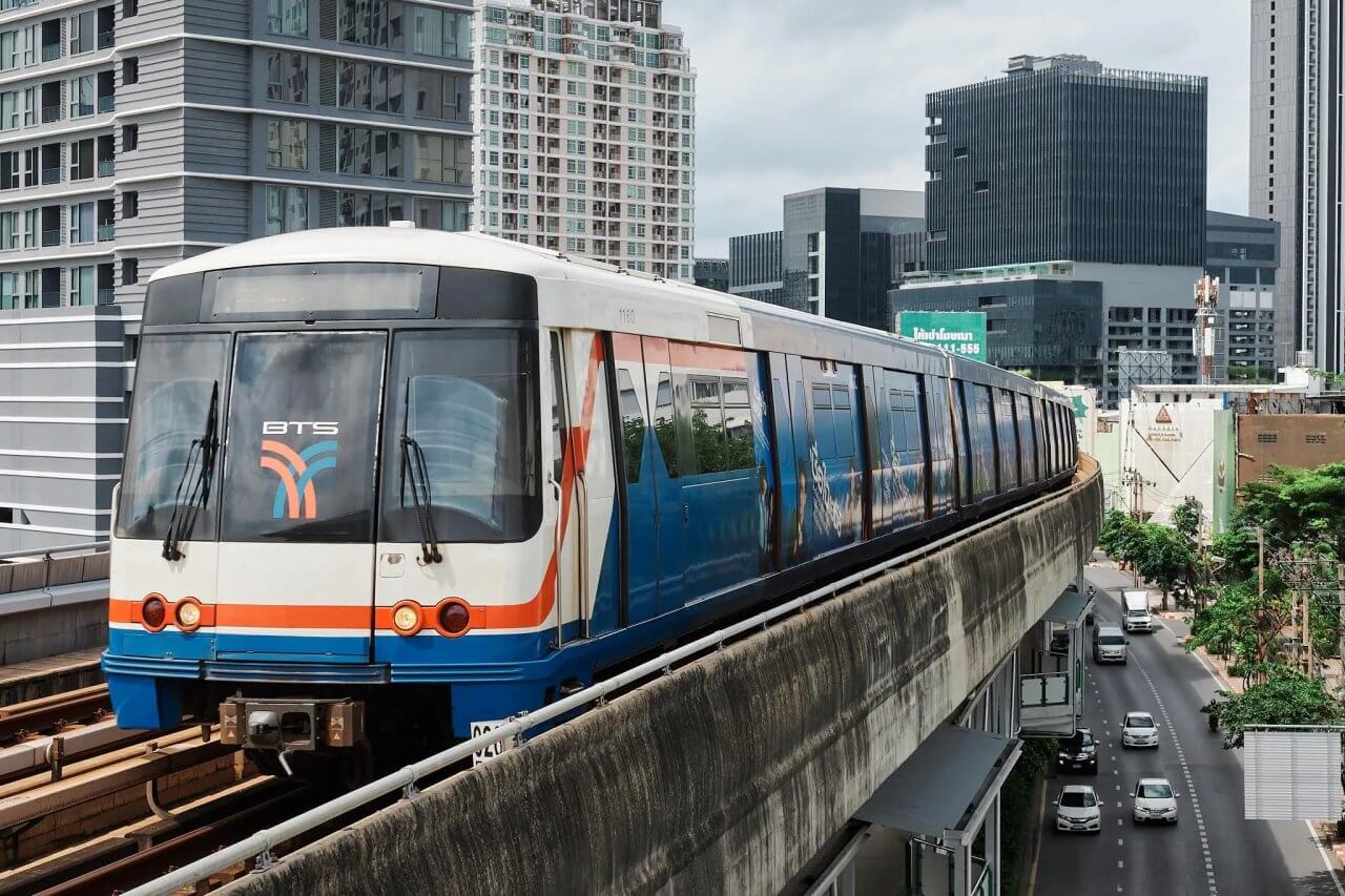 BTS Skytrain traveling on an elevated track amidst modern buildings in a bustling urban setting of Bangkok, Thailand.