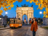 Kannaya Nareswari enjoying an autumn evening stroll by Milan's historic arch, with vibrant yellow leaves and a vintage tram in view.