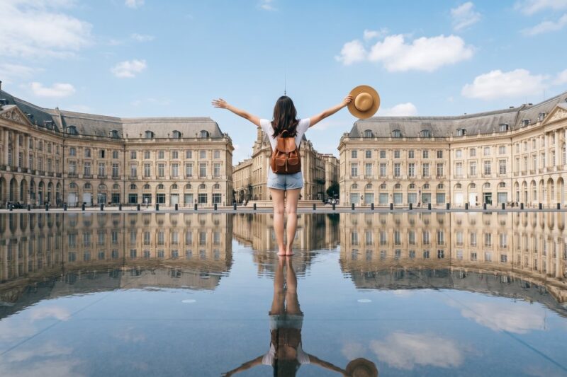 Girl enjoying a sunlit moment at Bordeaux's Place de la Bourse, reflecting on the water's surface with arms wide open.