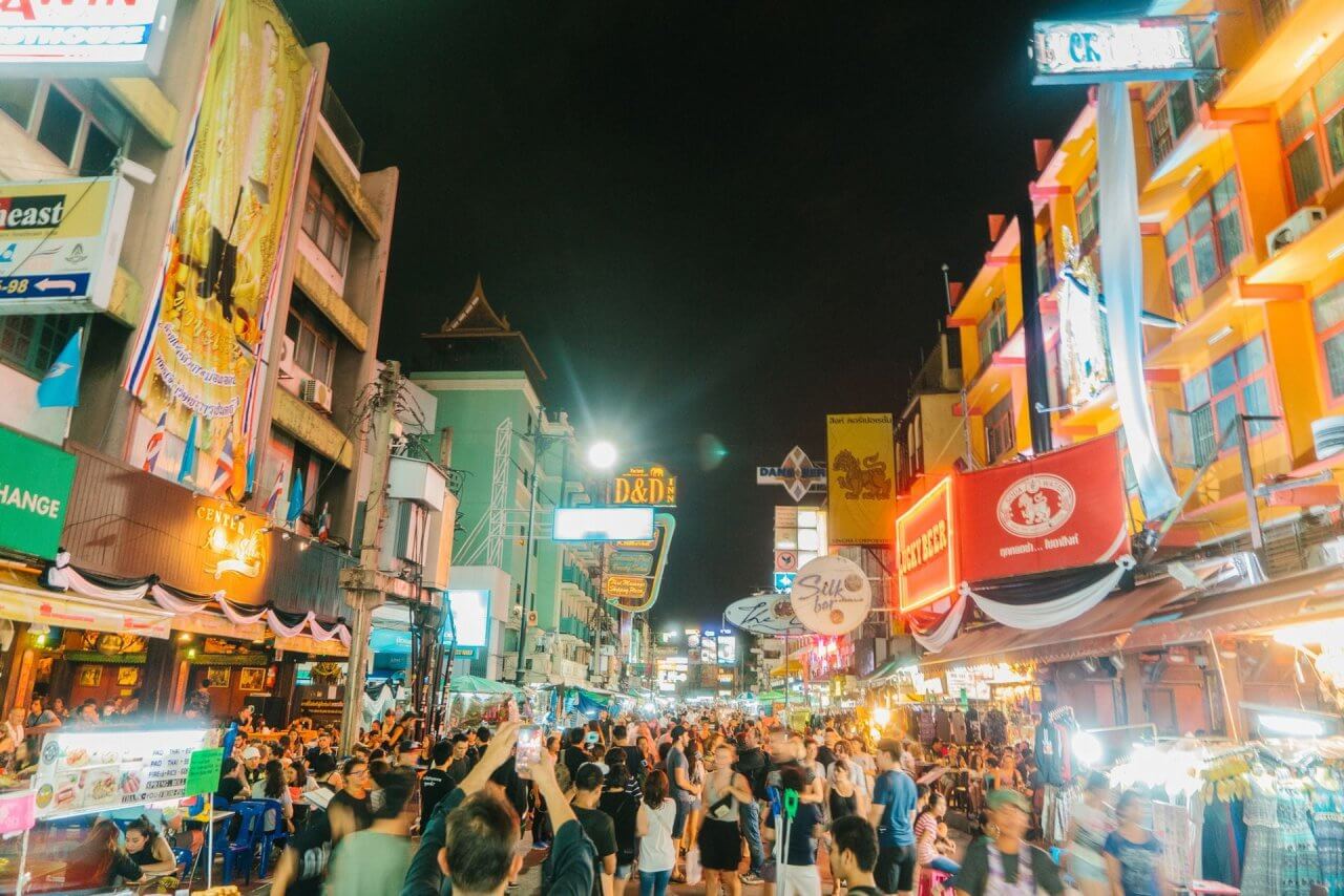 Bustling nightlife scene on a vibrant street filled with people, colorful neon signs, and local eateries in Bangkok.