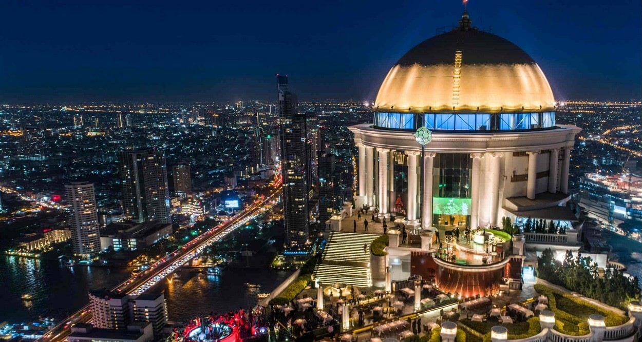 Nighttime cityscape featuring a luxurious rooftop venue with stunning views of Bangkok's skyline and the Chao Phraya River.