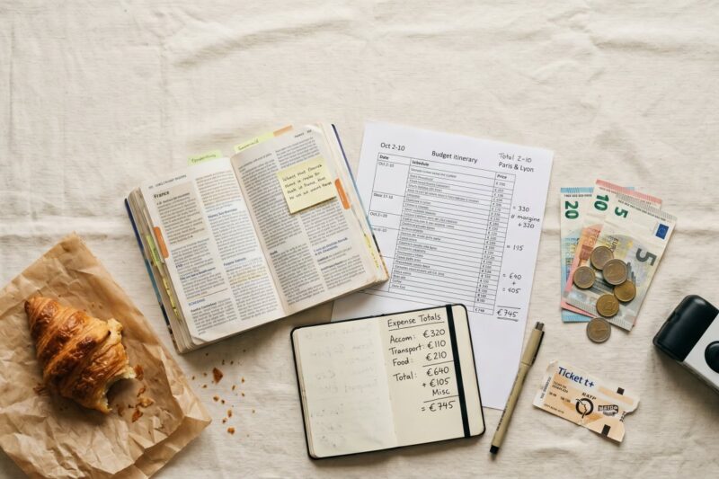 Planning a trip, this flat lay features a budget itinerary, travel expenses, a guidebook, a croissant, and a camera.