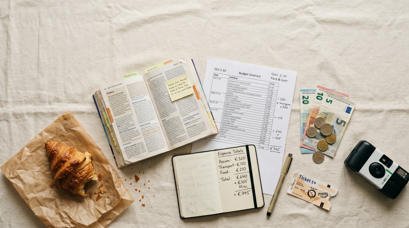 Planning a trip, this flat lay features a budget itinerary, travel expenses, a guidebook, a croissant, and a camera.