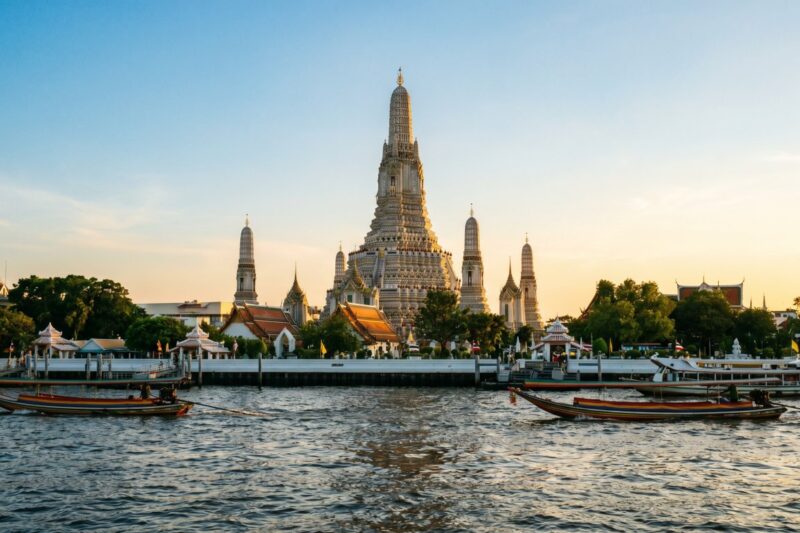 Wat Arun stands majestically along the riverbank, with traditional boats gliding past at sunset in Bangkok, Thailand.