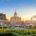 Golden spires of Wat Benchamabophit in Bangkok surrounded by vibrant gardens and golden marigolds under a sunset sky.