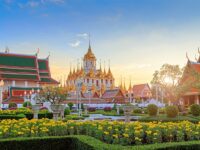 Golden spires of Wat Benchamabophit in Bangkok surrounded by vibrant gardens and golden marigolds under a sunset sky.