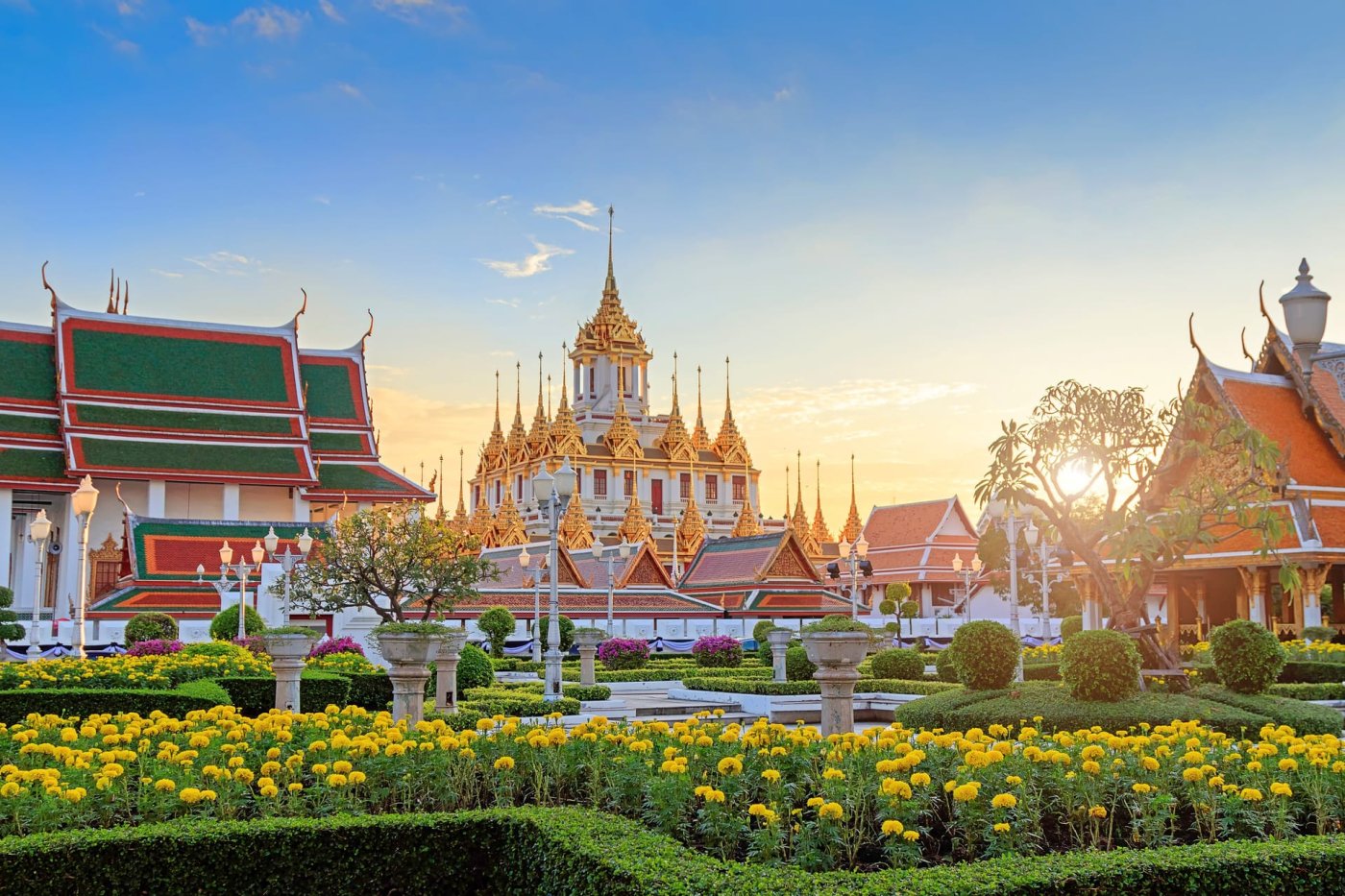 Golden spires of Wat Benchamabophit in Bangkok surrounded by vibrant gardens and golden marigolds under a sunset sky.