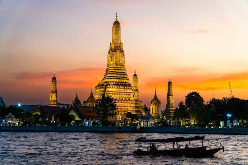 Wat Arun illuminated at sunset with boats on the Chao Phraya River, showcasing Bangkok's stunning skyline and rich culture.