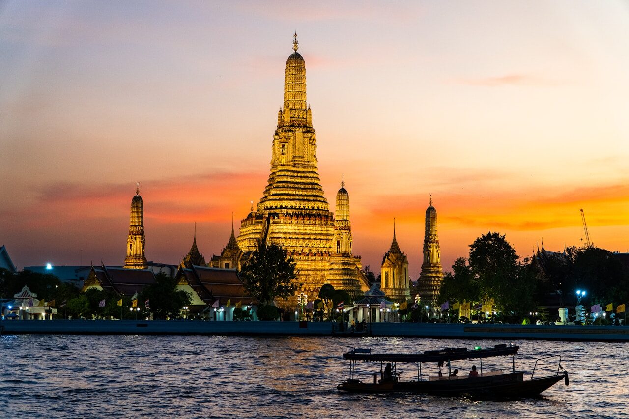 Wat Arun illuminated at sunset with boats on the Chao Phraya River, showcasing Bangkok's stunning skyline and rich culture.
